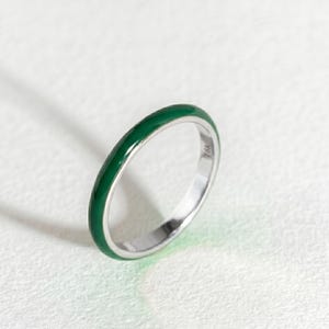 May include: A thin, circular ring featuring a smooth, vibrant green enamel inlay on the outer surface. The band is constructed from a metallic material with a polished silver-tone finish. The interior of the band is clearly stamped with the text 18K.