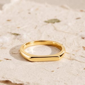 Può includere: Un anello color oro lucido caratterizzato da un design a barra rettangolare piatta e liscia sulla parte anteriore. La fascia è di larghezza uniforme e presenta una finitura metallica molto brillante che riflette la luce.
