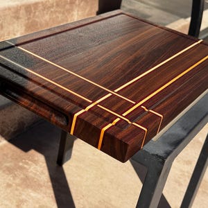 May include: A dark brown wooden chopping board with a juice groove. It has inlaid golden stripes that intersect at the corners. The board is set on a black metal frame.