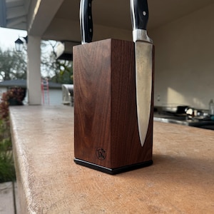May include: A dark brown wooden knife block with two knives. The block is rectangular with a black base and a logo on the front. The knives have black handles and silver blades.