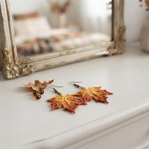 May include: A pair of drop earrings shaped like maple leaves, crocheted with variegated thread in shades of yellow, orange, and red. The earrings feature dark metal hooks and are displayed alongside a small, dried brown oak leaf and two beads.