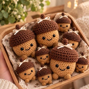 Puede incluir: Una colección de nueve bellotas de ganchillo en una bandeja de madera. Cada bellota tiene un sombrero texturizado de color marrón oscuro, un cuerpo color canela, dos ojos de cuentas negras, una sonrisa bordada y un lazo color crema.