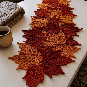 May include: A crochet table runner composed of interconnected maple leaf motifs in shades of deep red, burnt orange, and golden yellow. The runner features a detailed, open-work stitch pattern that mimics the veins of autumn leaves.