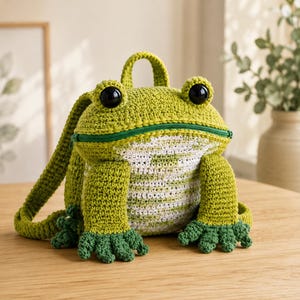 May include: A crochet backpack shaped like a frog in shades of lime green and white. The bag features large black plastic eyes, a green zippered mouth opening, and dark green webbed feet at the base. It includes a top handle and shoulder straps.