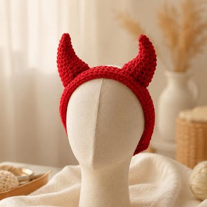 May include: A bright red crochet headband featuring two pointed, horn-shaped protrusions extending upward from the top. The headband is constructed with a thick, textured yarn stitch pattern and is displayed on a neutral-colored mannequin head.