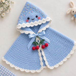 May include: A light blue hooded crochet cape featuring a smiling face on the hood with embroidered colorful sprinkles, a white scalloped trim, a light blue satin ribbon bow, and two pairs of small red crocheted cherries hanging from the center.