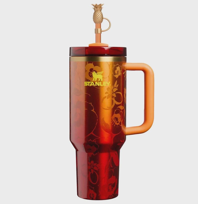 May include: A tall, cylindrical Stanley tumbler featuring a gradient red and orange finish with a floral pattern. It has a sturdy orange side handle, a dark red lid, and a straw topped with a small, gold-colored pineapple-shaped accessory.
