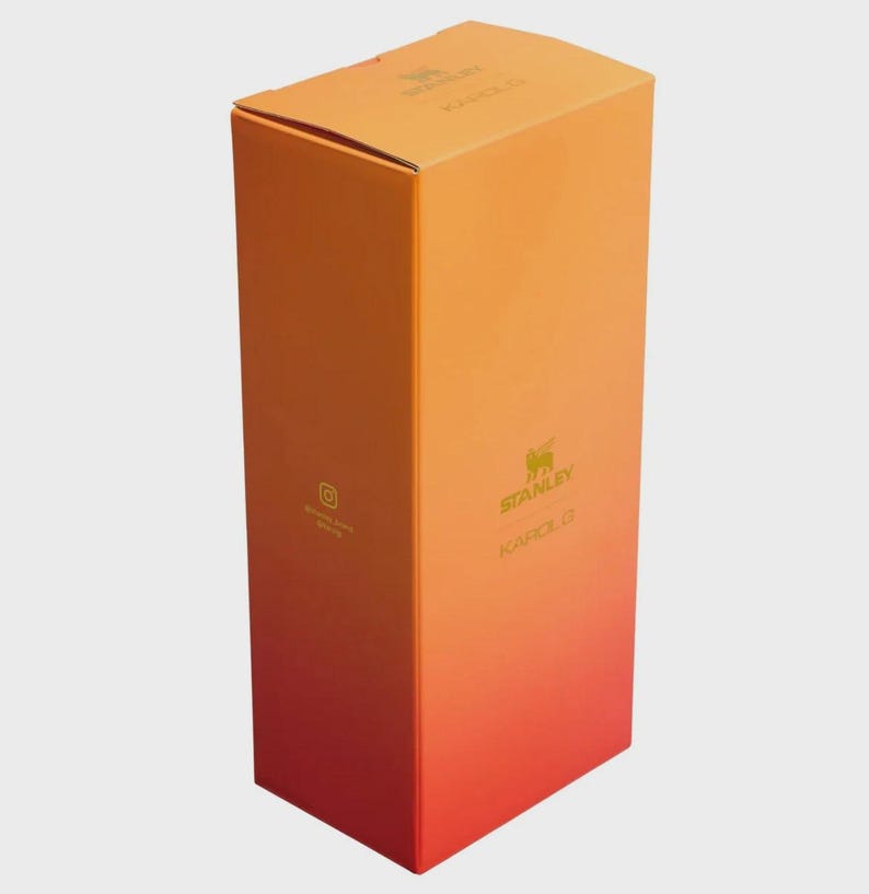 May include: A tall, rectangular cardboard product box featuring a vibrant orange-to-red gradient color scheme. The front displays the Stanley logo and the text KAROL G in gold lettering, with a small Instagram icon printed on the side panel.