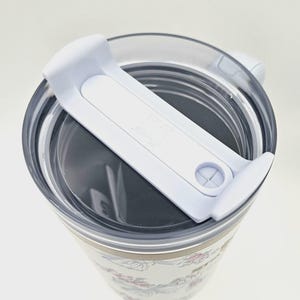 May include: A white tumbler with a floral pattern featuring pink and blue flowers and green leaves. The tumbler has a clear plastic lid with a white sliding closure mechanism on top. The design includes a small logo embossed on the white slider.