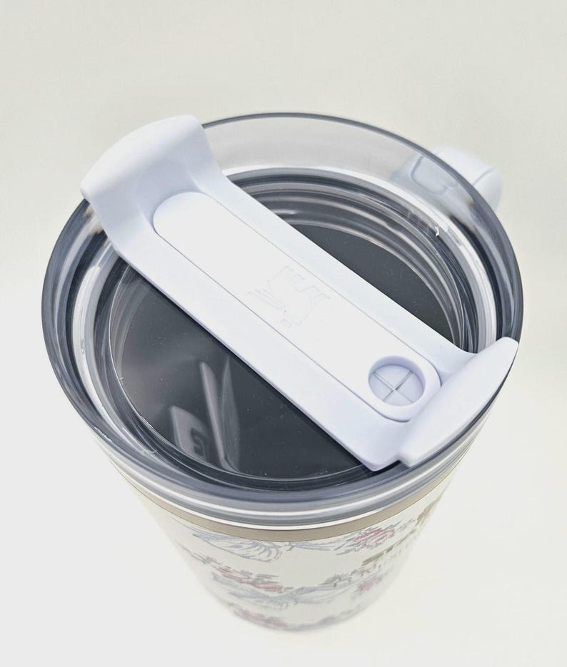 May include: A white tumbler with a floral pattern featuring pink and blue flowers and green leaves. The tumbler has a clear plastic lid with a white sliding closure mechanism on top. The design includes a small logo embossed on the white slider.