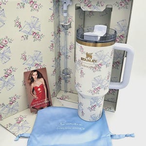 May include: A white Stanley tumbler with a floral and bow pattern, featuring a gold logo and the text LOVESHACKFANCY. The set includes a matching straw with a bow topper, a light blue drawstring pouch, and a card with the text LOVESHACKFANCY.