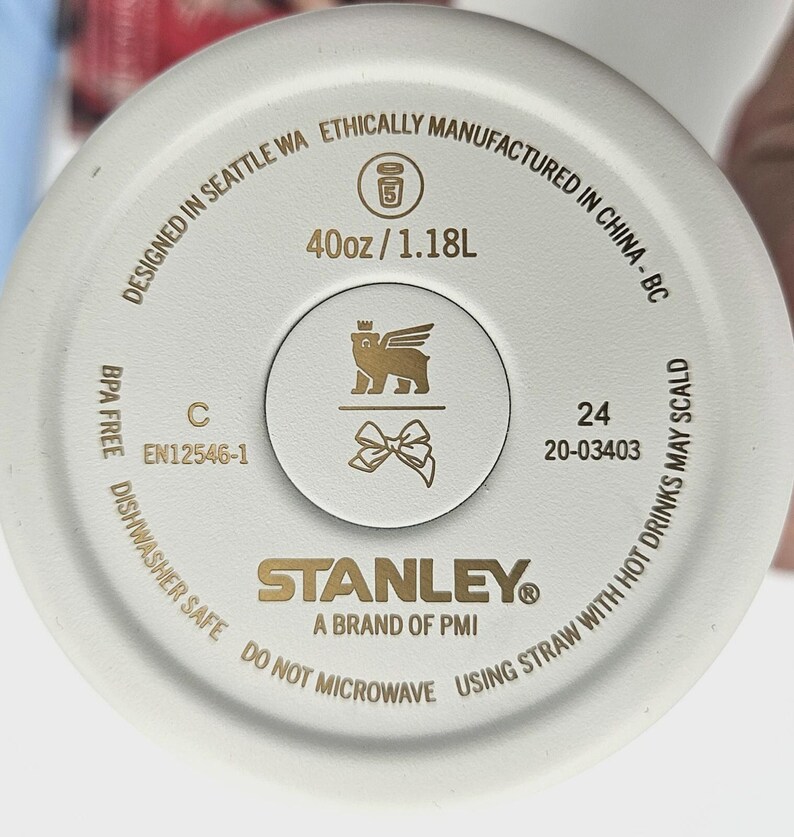 May include: The circular base of a cream-colored tumbler features gold text and icons. It displays '40oz / 1.18L', 'STANLEY A BRAND OF PMI', 'DESIGNED IN SEATTLE WA ETHICALLY MANUFACTURED IN CHINA', 'BPA FREE', 'DISHWASHER SAFE', and a bear with wings icon.