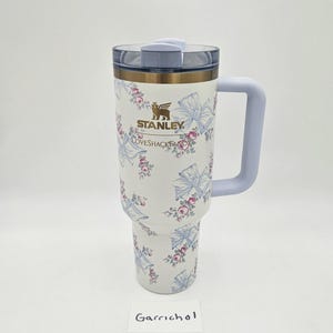 May include: A white Stanley LoveShackFancy tumbler with a floral and blue ribbon pattern, featuring a gold logo, a light blue handle, and a clear lid. The tumbler has a tapered base and a cylindrical body with a metallic gold rim near the top.