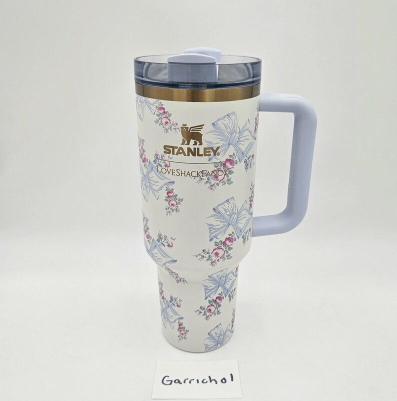 May include: A white Stanley LoveShackFancy tumbler with a floral and blue ribbon pattern, featuring a gold logo, a light blue handle, and a clear lid. The tumbler has a tapered base and a cylindrical body with a metallic gold rim near the top.