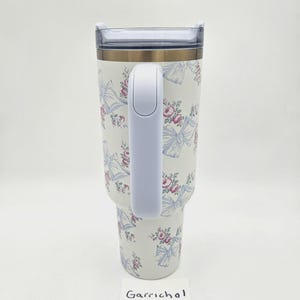 May include: A white insulated tumbler with a handle, featuring a repeating pattern of light blue bows and pink floral clusters. The tumbler has a clear plastic lid and a metallic rim. The word Garrichol is printed on a small white card at the base.