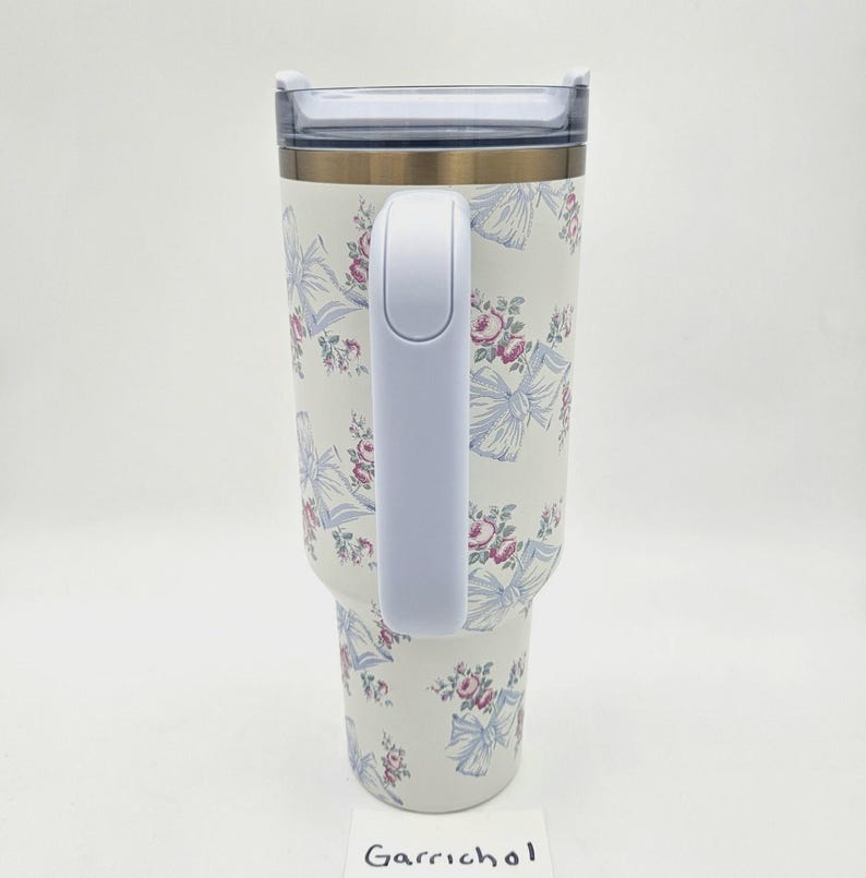 May include: A white insulated tumbler with a handle, featuring a repeating pattern of light blue bows and pink floral clusters. The tumbler has a clear plastic lid and a metallic rim. The word Garrichol is printed on a small white card at the base.