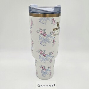 May include: A white Stanley tumbler featuring a repeating pattern of pink roses and light blue bows. The cup has a clear plastic lid with a white rotating closure. The Stanley logo and the text LOVESHACKFANCY are printed in gold on the front.