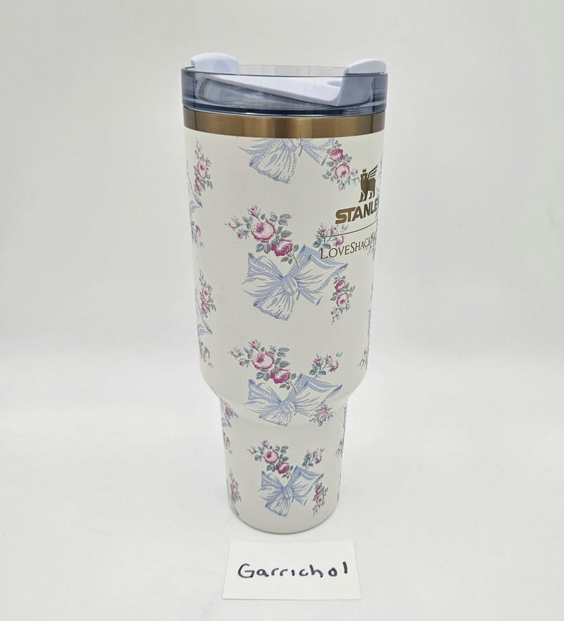 May include: A white Stanley tumbler featuring a repeating pattern of pink roses and light blue bows. The cup has a clear plastic lid with a white rotating closure. The Stanley logo and the text LOVESHACKFANCY are printed in gold on the front.