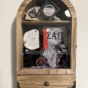 May include: A wooden shadow box with a glass front, displaying memorabilia. Inside are items like a medal, a red and black ribbon, and a graduation tassel. The box features the Purdue University logo and the text "Honoring Those Who Serve."