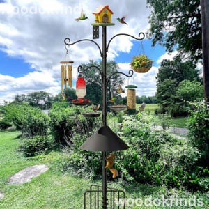 May include: A black metal bird feeder station with multiple hanging feeders, a birdhouse, and a squirrel baffle. The feeders include a hummingbird feeder, a seed feeder, and a hanging basket of flowers. The station is in a garden setting.