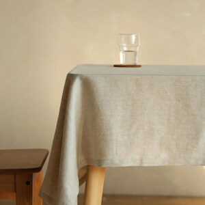 Op de afbeelding: Een lichtgrijze linnen tafelkleed over een houten tafel. Een glas water staat op een kleine onderzetter. Een houten stoel is gedeeltelijk zichtbaar. De scène wordt verlicht door zacht, natuurlijk licht.