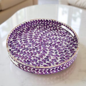 Op de afbeelding: Ronde decoratieve schaal met een witte achtergrond en een patroon van paarse bladvormen. De rand van de schaal is versierd met kleine, mozaïekachtige tegels. De schaal heeft een handvat.