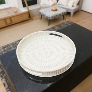 Puede incluir: Una bandeja decorativa redonda, de color blanco roto, con un diseño geométrico. La bandeja tiene dos asas integradas y está colocada sobre una superficie oscura. El diseño de la bandeja presenta un patrón radial de formas repetidas.