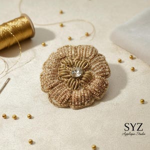 Puede incluir: Un broche artesanal en forma de flor de color dorado con un cristal central. El broche está rodeado de hilo y cuentas doradas. También se ven un carrete de hilo dorado, una aguja y un trozo de tela.