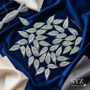 以下が含まれることがあります： 深い青色のベルベットの表面に配置された、銀色の葉のアップリケのコレクション。葉は、テクスチャ加工されたデザインで複雑に詳細に描かれています。刺繍糸と針、生地の見本も表示されています。画像には「SYZ Applique Studio」の文字が含まれています。