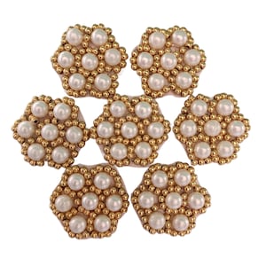 Peut inclure: Sept embellissements floraux décoratifs. Chacun a une base en tissu beige clair, un anneau de petites perles dorées et un groupe de fausses perles blanches. Les embellissements sont disposés en cercle sur fond blanc.