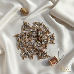 May include: Leaf-shaped appliques with gold, silver, and brown embroidery, adorned with clear rhinestones. The appliques are arranged on a white satin surface, with spools of thread and a needle. The appliques are from SYZ Applique Studio.