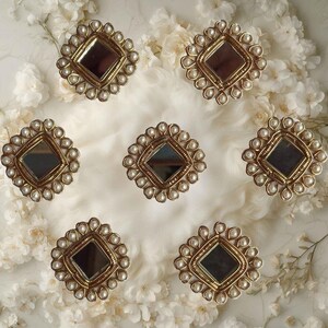 May include: Seven decorative, gold-toned, diamond-shaped objects with a central mirror and a border of small, white pearls. The objects are arranged on a white, textured surface, surrounded by white flowers.