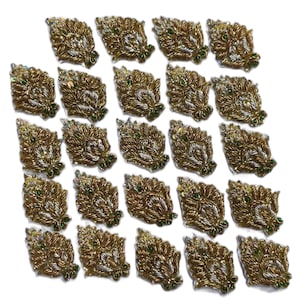 May include: A collection of gold-coloured, leaf-shaped embroidered appliques. Each applique features intricate detailing with gold thread, silver accents, and small green beads. The appliques are arranged on a white surface.