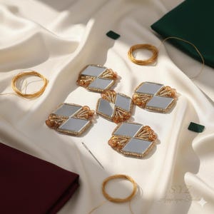 May include: Five decorative, mirrored embellishments with gold trim and orange bead accents are arranged on a white satin surface. Gold thread, a needle, and fabric squares in burgundy and green are also present. The image is well-lit, highlighting the details.