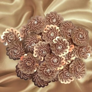 Puede incluir: Un conjunto de adornos florales de lentejuelas en oro rosa. Cada flor tiene un brillante central transparente, rodeado de hilo metálico en espiral y lentejuelas. El fondo es de tela de satén.