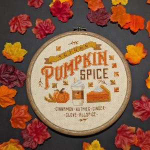 以下が含まれることがあります： オレンジとクリーム色の「Pumpkin Spice」の文字が入ったクロスステッチ刺繍枠。デザインにはカボチャ、コーヒー、パイのスライスが含まれています。「Cinnamon, Nutmeg, Ginger, Clove, Allspice」の文字も含まれています。枠はカラフルな秋の葉で囲まれています。