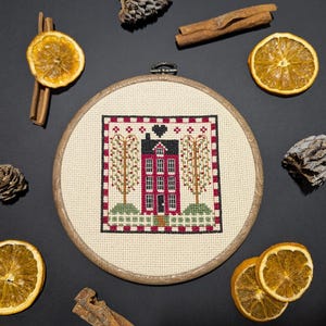 May include: A cross-stitch embroidery hoop featuring a red house with a black roof, flanked by trees. The design is framed with a red and white border. Dried orange slices, cinnamon sticks, and pine cones surround the hoop on a dark surface.