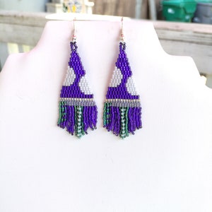 May include: A pair of beaded earrings in a triangular shape. The earrings have a purple background with white and grey circular patterns. The bottom fringe is a mix of green, purple, and grey beads. The earrings are displayed on a white bust.