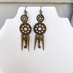 May include: A pair of bronze-toned dangle earrings featuring a vertical arrangement of two circular gears, a central decorative key charm, and two pointed spikes hanging from the bottom of the larger gear, suspended by standard earring hooks.