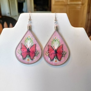 May include: A pair of pink teardrop-shaped earrings with a silver wire frame and a butterfly design. The butterfly is pink and has black markings.