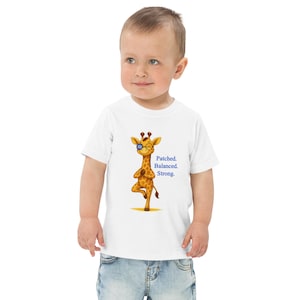 Pode incluir: Uma camiseta branca para crianças pequenas com uma girafa de desenho animado usando óculos e equilibrando-se em uma perna. A girafa é marrom e amarela. O texto "Patched. Balanced. Strong." é impresso em azul.