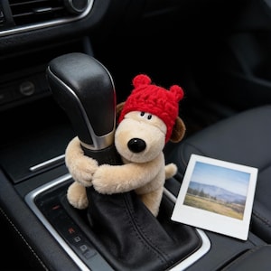 Pode incluir: Um brinquedo de pelúcia de cachorro usando um chapéu de malha vermelho, abraçando a alavanca de câmbio de um carro. O cachorro é bege com orelhas marrons e um nariz preto. Uma pequena foto de uma paisagem está ao lado do cachorro.