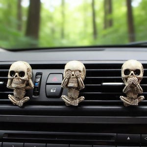 May include: Three beige skull car air fresheners in a "see no evil, hear no evil, speak no evil" pose. The skulls are seated with their hands covering their eyes, ears, and mouth. The air fresheners are on a black car dashboard with a blurred green background.