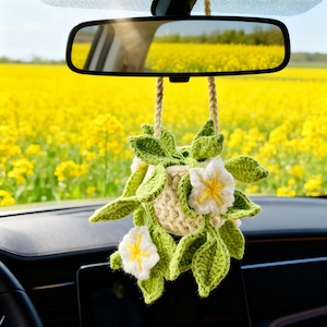 May include: A handmade crochet car charm featuring a cream-colored basket with white flowers and green leaves. The charm hangs from the rearview mirror, with a field of yellow flowers reflected in the mirror.