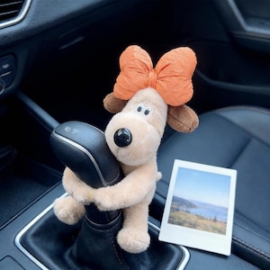 May include: A plush dog toy with a large orange bow on its head, hugging a car gear shift. The dog is light brown with brown ears and a black nose. A small photo of a landscape is next to the dog.