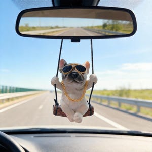 Könnte beinhalten: Ein Auto-Accessoire mit einer weißen und grauen Bulldogge, die eine Sonnenbrille und eine goldene Kette trägt und auf einer Holzschaukel sitzt. Das Accessoire hängt am Rückspiegel, mit einer Autobahn im Hintergrund.