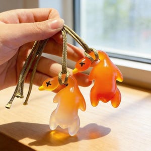 May include: Three orange and white duck-shaped pendants with black cross details on their beaks, suspended from olive green cords. The pendants have a glossy, translucent appearance. The background is blurred, suggesting a window and a wooden surface.