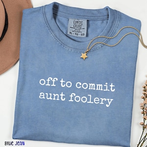 May include: A blue jean coloured t-shirt with a crew neckline and a Comfort Colors brand label at the inner collar. The front of the shirt features the white text "off to commit aunt foolery" in a typewriter font style. A gold star pendant necklace.