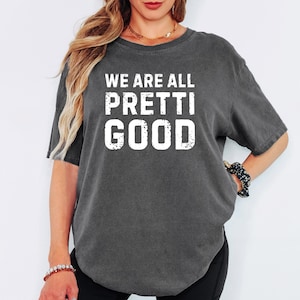 May include: Dark gray t-shirt with the text "WE ARE ALL PRETTI GOOD" in white, distressed lettering. The shirt has short sleeves and a relaxed fit. The overall style is casual and comfortable.