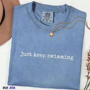 May include: A blue jean colored t-shirt with the phrase "just keep swimming" printed in white. A gold star pendant necklace rests on the shirt. The shirt has a Comfort Colors tag.